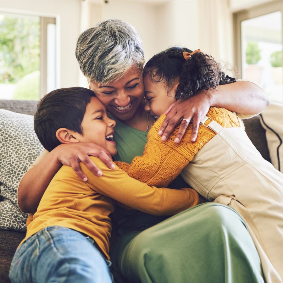 An older woman is sitting on the couch hugging two kids. All three are smiling.