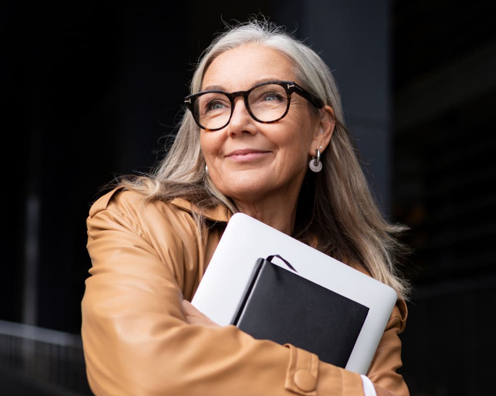 Older woman with glasses outside smiling holding a laptop and notebook.