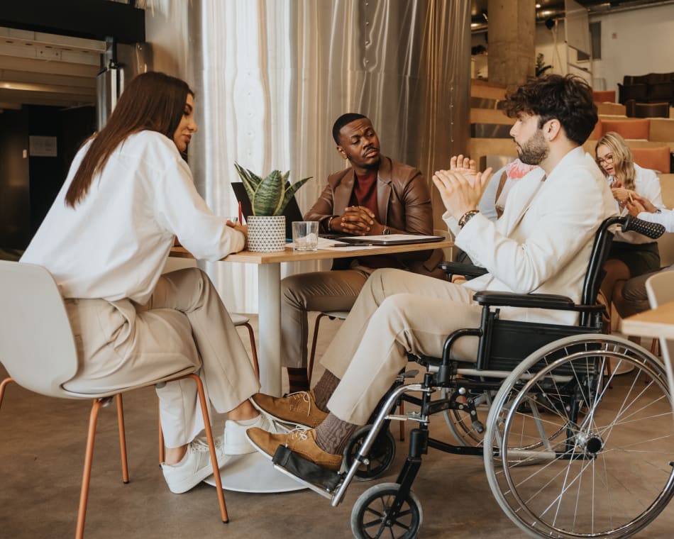 Group of young adults sitting around a table talking. One man is sitting in a wheelchair.